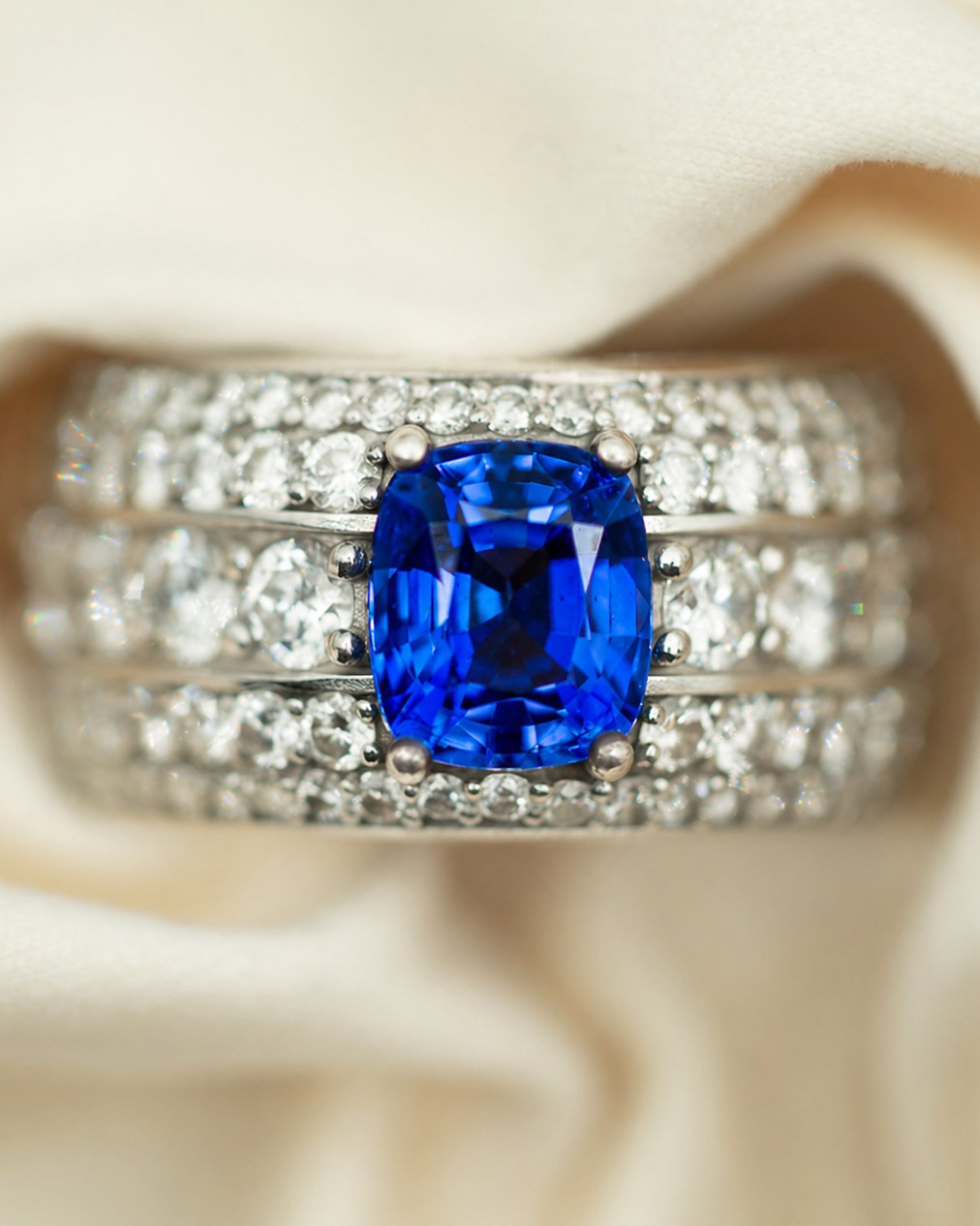 Cushion-cut blue sapphire centre stone in a triple-row diamond pavé band, set in white gold, on cream silk